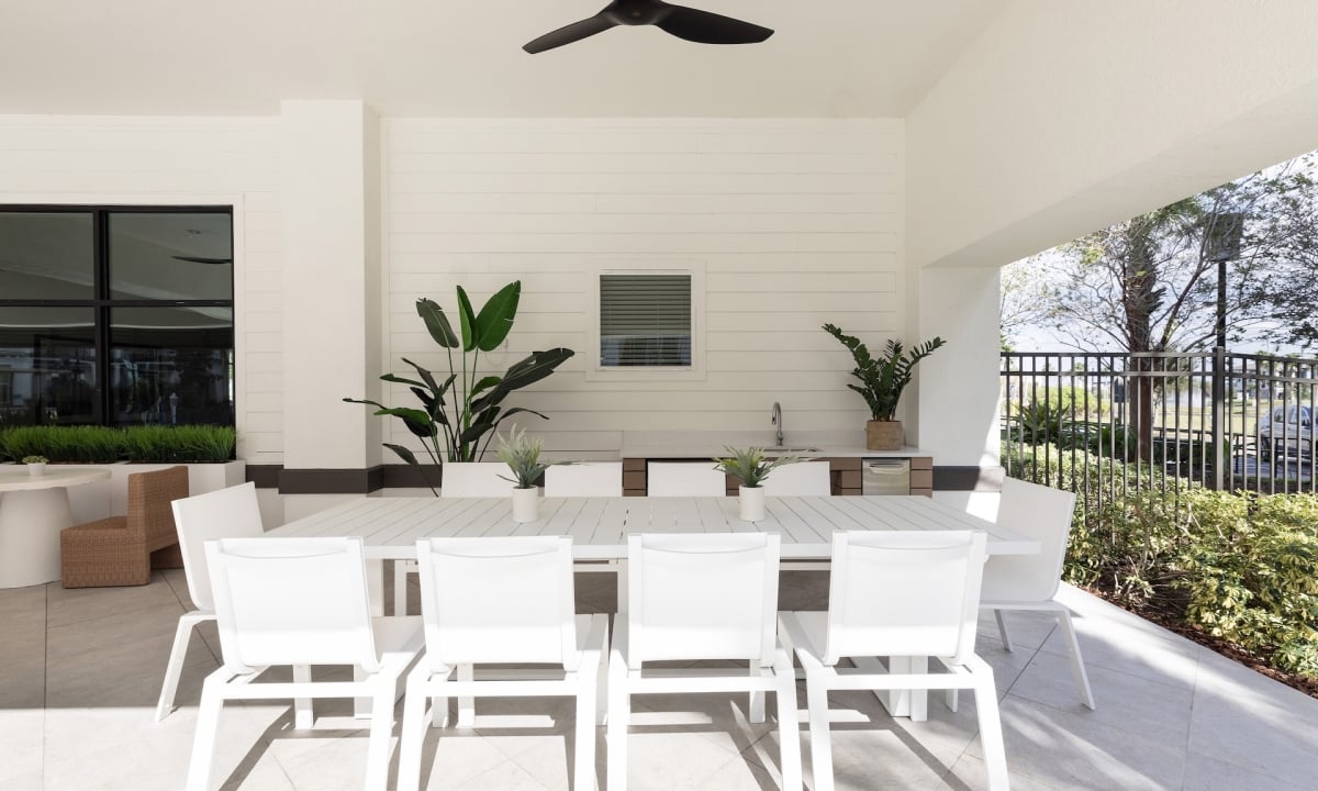 A white outdoor dining area with a large table and chairs, plants, and a ceiling fan.