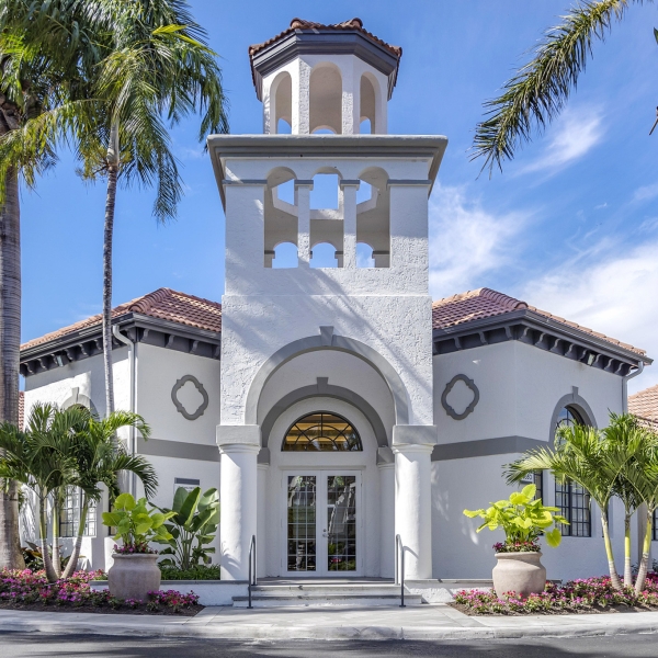 A white stucco building with a tower and arched entrance, surrounded by palm trees and flowering plants.